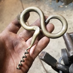 May include: A hand holds a gold-colored metal corkscrew with a unique, twisted handle. The corkscrew has a spiral design and is made of metal.