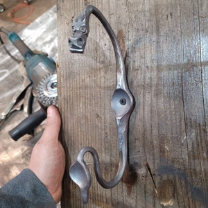May include: A metal hook shaped like a dragon with a decorative head and a single screw. The hook is mounted on a wooden surface.