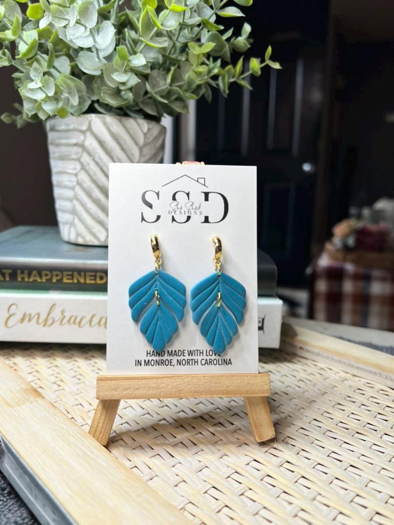 May include: Teal blue leaf-shaped earrings with gold hoops. The earrings are displayed on a white card with the text "SSD Ae Aed DESIGNS" and "HAND MADE WITH LOVE IN MONROE, NORTH CAROLINA".
