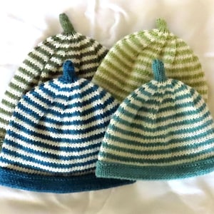 Pode incluir: Quatro gorros de malha com riscas horizontais em várias cores, incluindo verde escuro, verde claro, azul e azul-petróleo. Cada gorro tem uma aba de cor sólida e um pequeno topo pontiagudo. A textura da malha é visível em cada peça.