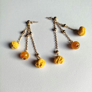 May include: A pair of gold-toned dangle earrings. Each earring features a post, small gold beads, a chain, a yellow fabric flower, and a round, orange-yellow bead. The earrings are set against a white background.