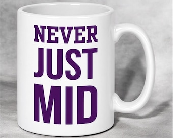 May include: A white ceramic mug featuring the text NEVER JUST MID printed in bold purple capital letters on the front. The mug has a smooth finish, a cylindrical body, and a curved handle on the right side for holding the vessel.