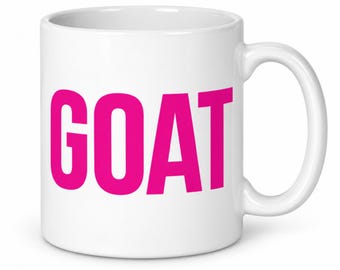 May include: A white ceramic mug featuring the word GOAT printed in bold, bright pink capital letters on the front. The mug has a smooth, glossy finish, a cylindrical body, and a curved handle on the right side for holding the vessel.