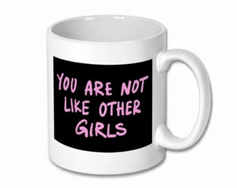 May include: A white ceramic mug featuring a black rectangular graphic on the front. The text "YOU ARE NOT LIKE OTHER GIRLS" is printed in a light pink, handwritten-style font centered within the black rectangle on the mug's exterior.