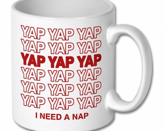 May include: A white ceramic mug featuring the repeated red text YAP YAP YAP in a grid pattern, with the phrase I NEED A NAP printed in red at the bottom. The mug has a standard cylindrical shape with a curved handle on the right side.