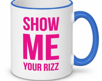 May include: A white ceramic mug with a bright blue rim and handle. The front of the mug features the text SHOW ME YOUR RIZZ printed in bold, vibrant pink capital letters, with the word SHOW ME stacked above the word YOUR RIZZ.