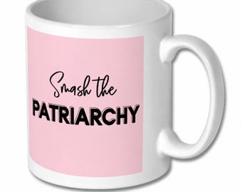 May include: A white ceramic mug featuring a light pink rectangular panel on the front. The text Smash the PATRIARCHY is printed in black, with the word Smash in a cursive script and PATRIARCHY in a bold, block-style sans-serif font.