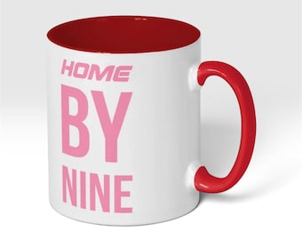 May include: A white ceramic mug with a red interior and a red handle. The exterior features the text HOME BY NINE printed in a bold, light pink sans-serif font. The mug has a cylindrical shape with a smooth finish and a sturdy side handle.