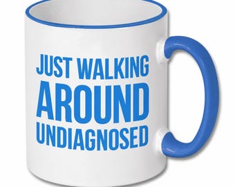 May include: A white ceramic mug with a blue rim and a blue handle. The front of the mug features the text "JUST WALKING AROUND UNDIAGNOSED" printed in bold, blue, sans-serif capital letters centered on the exterior surface of the mug.
