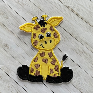 May include: A yellow and brown embroidered giraffe applique with black feet. The giraffe is sitting with its legs crossed and has a friendly expression.