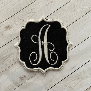 May include: A black and white embroidered patch with a white letter "A" in a decorative script font. The patch is surrounded by a white border and has a scalloped edge.