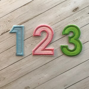 May include: Three embroidered fabric numbers: 1 in light blue, 2 in pink, and 3 in green. Each number has a white outline and is stitched onto a light-colored wooden surface. The numbers are likely for crafts or decorations.