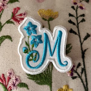 May include: A white embroidered patch featuring the letter M in bright blue thread, accented with two small blue flowers and green stems. The patch has a white border and is placed against a fabric background with floral embroidery.