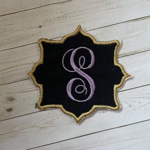 May include: Black embroidered patch with a gold border and a lavender script letter "S". The patch has a decorative, scalloped edge. The patch is on a light wood grain surface.