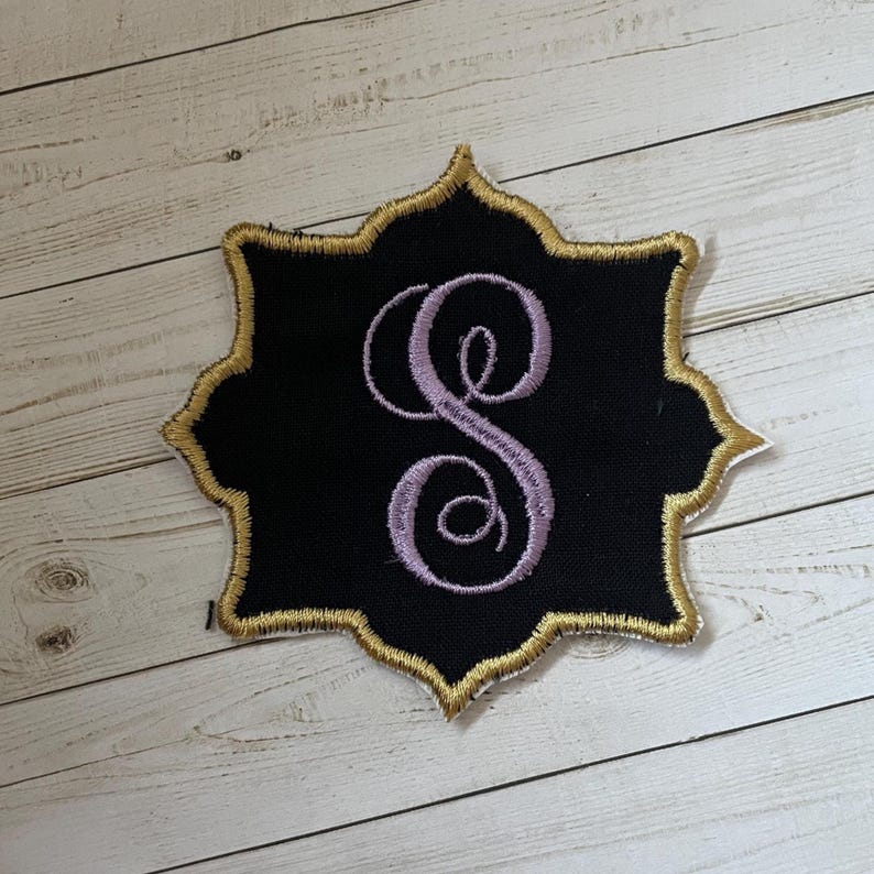 Peut inclure: &Eacute;cusson brod&eacute; noir avec une bordure dor&eacute;e et une lettre "S" en script lavande. Le patch a un bord d&eacute;coratif festonn&eacute;. Le patch est sur une surface de grain de bois clair.