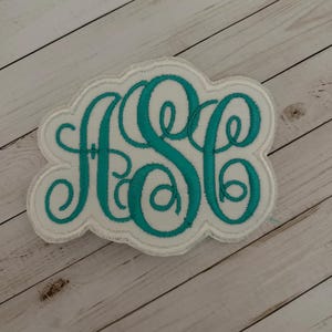 May include: A white fabric patch featuring an embroidered monogram with the letters ASC in a teal cursive script. The patch has a scalloped edge and a white border, with the teal thread creating a decorative, swirling design for the letters.