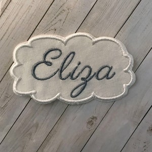 May include: White fabric cloud-shaped patch with the name "Eliza" embroidered in gray thread.
