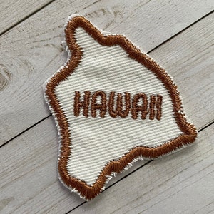 May include: A white fabric patch in the shape of the state of Hawaii with a brown embroidered outline and the word "HAWAII" embroidered in brown.