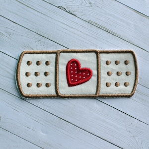 May include: A white fabric bandage with a brown stitched border and a red heart with white polka dots in the center.