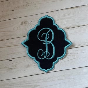 May include: Embroidered patch featuring the letter "B" in a teal script, set against a black background. The patch has a decorative teal border and a unique, ornate shape. The patch is likely for clothing or accessories.