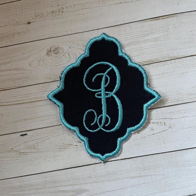May include: Embroidered patch featuring the letter "B" in a teal script, set against a black background. The patch has a decorative teal border and a unique, ornate shape. The patch is likely for clothing or accessories.