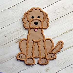 May include: A brown embroidered patch of a cartoon poodle dog with a pink tongue sticking out. The dog is sitting with its legs crossed and has a brown nose and black eyes.