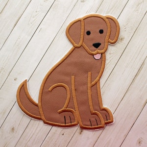 May include: A brown embroidered patch in the shape of a sitting dog. The dog has a white outline, black eyes, a black nose, and a pink tongue. The patch is on a light wood-grain background.