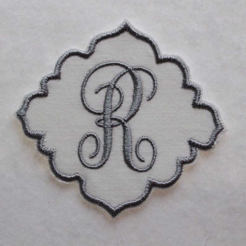 May include: A gray embroidered monogram letter "R" on a white fabric patch. The patch has a scalloped edge and is suitable for sewing onto clothing or other fabric items. The embroidery is detailed and elegant.