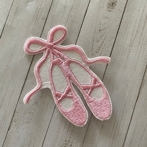 May include: Pink felt ballerina shoe applique with a white outline and a bow. The applique is perfect for adding a touch of whimsy to crafts and projects.