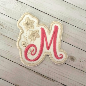 May include: An embroidered fabric patch featuring a large, pink, cursive letter M. The letter is surrounded by a cream-colored border and includes two stitched cream-colored flowers on the left side, all set against a white fabric background.