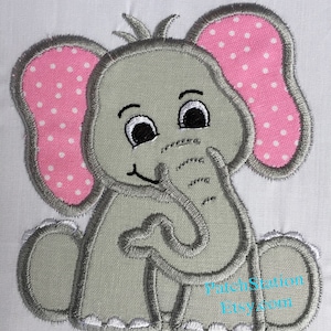 May include: Embroidered gray elephant applique with pink polka dot ears. The elephant is sitting with its trunk in its mouth and has a smiling face.