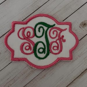 May include: A white fabric patch with a scalloped border embroidered in pink thread. The center features a monogram with the letters S, J, and F stitched in a decorative script font. The letters S and F are in pink, while the letter J is in green.
