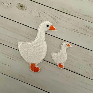 May include: Two white goose embroidered patches with orange beaks and feet. The larger goose is standing upright, while the smaller goose is standing with its head turned to the side.