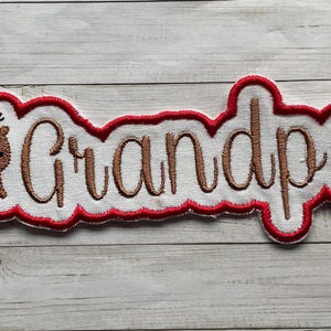 May include: Embroidered patch with the word "Grandpa" in brown script. A cartoon reindeer with a red scarf is on the left. The patch has a white background with a red scalloped border. Great for Christmas gifts.