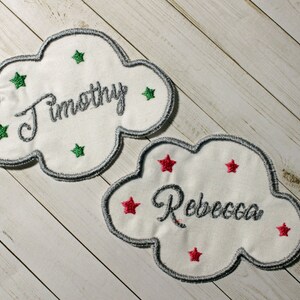 Pu&ograve; includere: Due toppe di tessuto bianco a forma di nuvola con nomi ricamati, "Timothy" e "Rebecca", e stelle verdi e rosse.