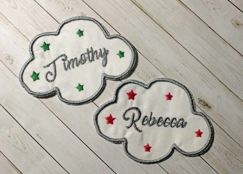 Pu&ograve; includere: Due toppe di tessuto bianco a forma di nuvola con nomi ricamati, "Timothy" e "Rebecca", e stelle verdi e rosse.