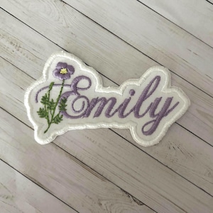 May include: A white fabric patch featuring the name Emily embroidered in a purple cursive script. To the left of the text, a small purple flower with a yellow center and green stem is embroidered onto the white background with a stitched border.