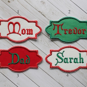 May include: Embroidered name tags in various colors. The tags feature the names "Mom", "Trevor", "Dad", and "Sarah" in decorative fonts. Each tag has a white or colored background with a contrasting border.