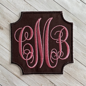 May include: A brown fabric patch with a pink embroidered monogram of the letters "CWB" in a script font.