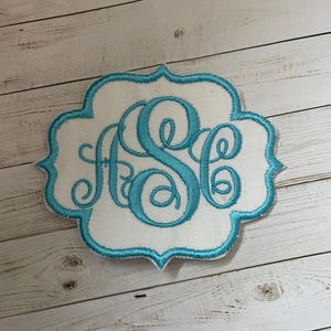 May include: Embroidered monogram patch featuring the letters "ASC" in a teal script font. The letters are stitched onto a white fabric, framed by a teal scalloped border. The patch is designed for personalization and customization.