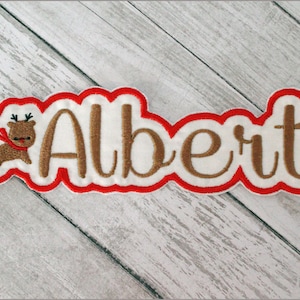 May include: Embroidered patch with the name "Albert" in brown script. The patch has a white background with a red border and a cartoon reindeer wearing a red scarf. The patch is designed for clothing or accessories.