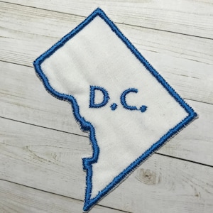 May include: A white fabric patch in the shape of the District of Columbia with a blue outline. The patch has the text "D.C." embroidered in blue.