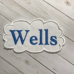 May include: White fabric patch with a blue embroidered word "Wells" in a cloud-shaped outline.