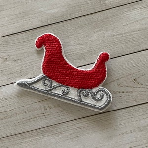 May include: Embroidered patch of a red and white Santa's sleigh with a gray base. The sleigh features detailed stitching and a classic design, perfect for holiday-themed crafts and decorations.
