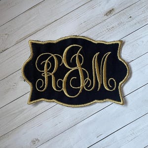 May include: Black embroidered patch with gold trim and gold cursive initials "RSM". The patch has a decorative, scalloped edge. The background is a light wood grain.