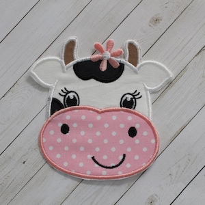 May include: A pink and white cow applique with a black nose and eyes. The cow has a pink flower on its head and a pink polka dot nose.