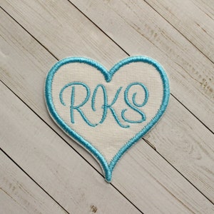 May include: A white heart-shaped patch with a blue outline and the monogram "RKS" embroidered in blue thread.