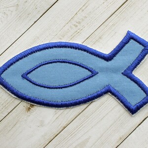 May include: A light blue embroidered fish patch with a dark blue outline. The patch is in the shape of a fish, with a detailed design. The patch is made of fabric and has a white backing.