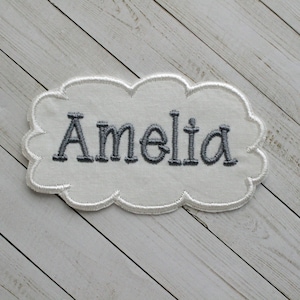 May include: White fabric cloud-shaped patch with gray embroidered lettering that reads "Amelia".