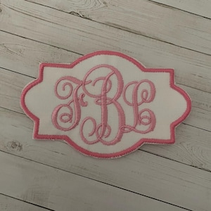 May include: A white fabric patch with a pink embroidered border. The patch features a pink monogram of the letters FBL in a decorative script. The patch has a unique, curved rectangular shape.